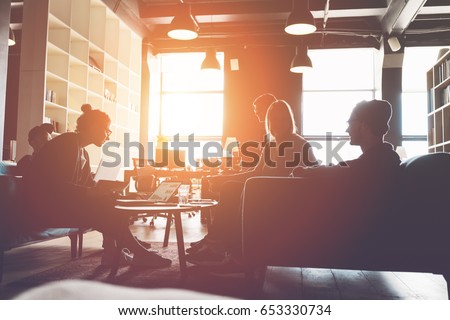 Silhouette of business group brainstorming new plan. Team meeting on the couch. Big open space office. Five people. Intentional sun glare