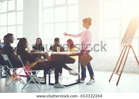 Male and female creative students listening explanation of female train manager during workshop sitting in office.Skilled couch conducting business seminar with young professionals.Advertising area Royalty-Free Stock Photo #653332264