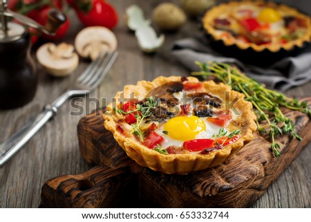 Mini tart with baked ham, pepper, mushrooms and quail eggs in creamy sauce on a wooden stand and old background. Selective focus.Top view.