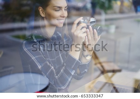 Attractive smiling female looking at vintage camera while making photo sitting at wooden table.Charming positive teenager woman spending leisure time in cafeteria with good mood and favorite hobby