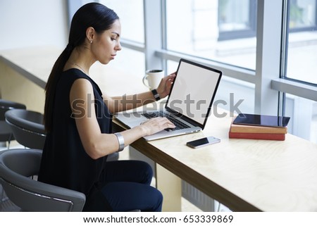 Concentrated attractive businesswoman banking online making money transfer using laptop computer with mock up screen connected to wifi, serious female entrepreneur doing remote work in cafe interior