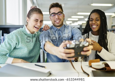 Positive male and female young designers making selfie on vintage camera during break sitting at desktop in office interior.Smiling friends taking picture and have fun enjoying recreation time
