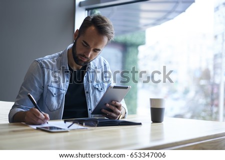 Handsome bearded student preparing for coursework project while browsing information in web pages and recording general concepts into paper.Concentrated author writing article while sitting in cafe
