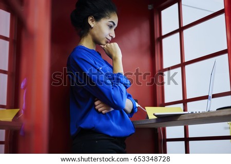 Thoughtful afro american student having online conversation via modern laptop device standing in isolated red cabine in coworking space.Concentrated female watching webinar on computer in wifi zone