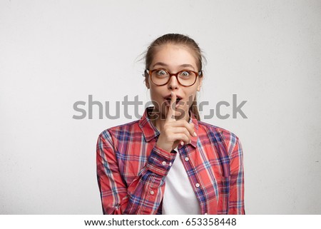Don't tell anyone! Attractive young European female wearing hair in ponytail, keeping index finger at her lips, hushing, asking to hold tongue and not unlock her secrets, having excited look