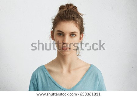 Portrait of young European female with healthy clean skin and blue eyes wearing casual top looking at camera with serious expression. Caucasian woman model with hair gathered in bunch posing indoors Royalty-Free Stock Photo #653361811