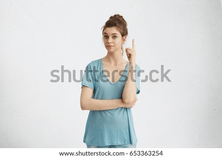 I have a great idea! Smart quick-witted pretty girl with hair bun keeping finger pointed upwards. Cute young Caucasian woman showing something above her head, making gesture with index finger