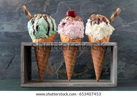 Pistachio, cherry and vanilla ice cream with topping in waffle cones in rustic wood holder over a slate background