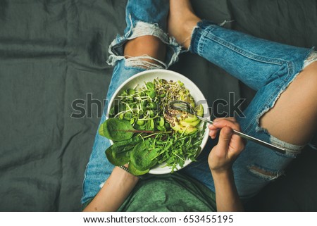 Green vegan breakfast meal in bowl with spinach, arugula, avocado, seeds and sprouts. Girl in jeans holding fork with knees and hands visible, top view, copy space. Clean eating, vegan food concept Royalty-Free Stock Photo #653455195