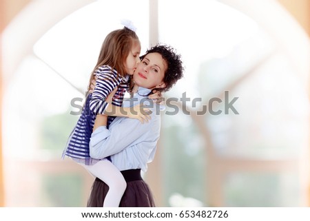 Happy young beautiful mother,lifted his little beloved daughter .And the girl kisses his mother on the cheek.In a room with a large semi-circular window.