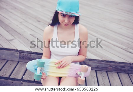 Skater girl with skateboard in hands.Young skateboarder girl sit in park.Pretty young brunette girl holding colorful mini skateboard in hands while sitting outdoor in good summer day.Teen skater girl