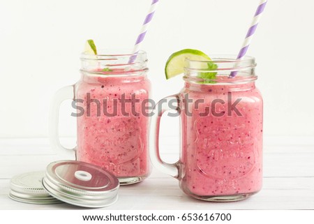 Smoothies with red berries and lime served in mason jars with colorful paper straws, white background.