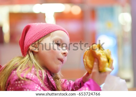 Adorable blonde girl in pink jacket and hat eat burger in fast food restaurant