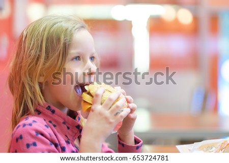 Adorable blonde cheerful girl in pink clothes eat double burger with beef and cheese in fast food restaurant