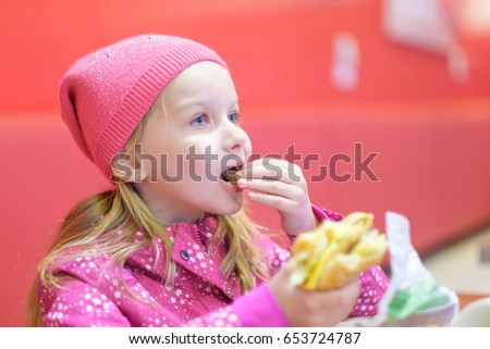 Adorable blonde girl in pink jacket and hat eat burger in fast food restaurant