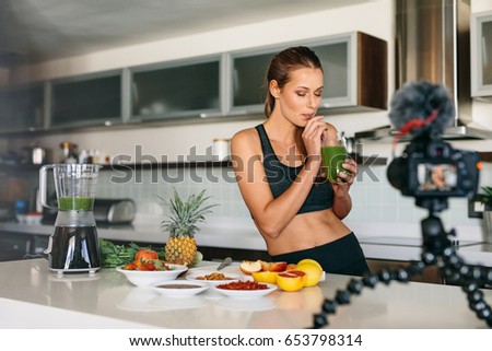 Young lady in kitchen drinking healthy vegetable juice while recording it on camera. Fruits and vegetables for breakfast on kitchen table.