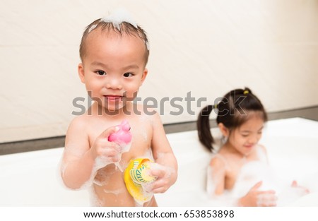 smiling happy little boy children holding duck toys and beautiful sweet girl playing foam bubble water in bathtub together on bathroom with selective focus photo.
