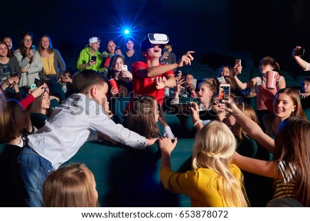 Young boy looking amazed and surprised pointing away with his finger wearing virtual reality glasses at the movie theatre his classmates looking at him copyspace film projector light beam technology.