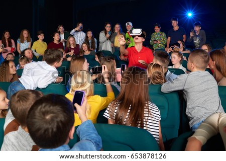 Group of kids taking pictures of their friend while he is wearing virtual reality goggles headset at the movie theatre 3D technology modern device gadget entertaining fun excitement concept.
