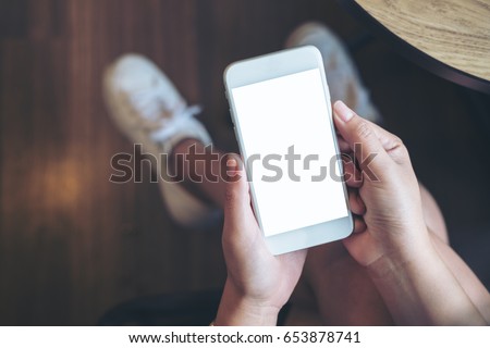 Mockup image of woman's hand holding white mobile phone with blank screen on thigh with wooden floor background in modern cafe