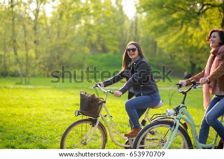Two young women ride bikes in the spring park. Gentle spring greens, yellow dandelions, clean air. Pleasure by nature and movements. Healthy lifestyle.