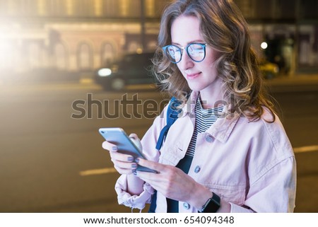 Hipster girl in glasses stands on night street and uses smartphone, touching screen with finger. Young woman is chatting,blogging,checking email, working.In background is road with cars in soft focus.