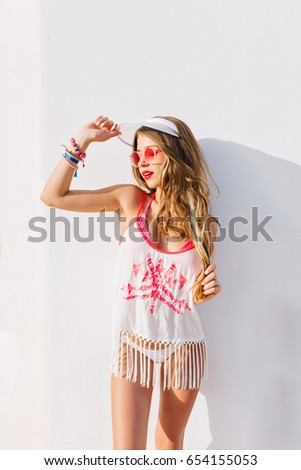 Graceful shapely woman with wonderful suntan wearing vintage fridge tank-top posing in front of wall. Charming long-haired lady in trendy hat and cute panties waving hand on white background