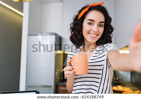 Cool brightful selfie portrait of attractive young woman with cut curly brunette hair smiling to camera with cup of tea in kitchen of modern apartment. Having fun, true positive emotions