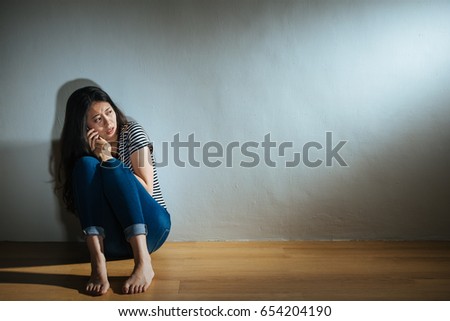 unhappy young woman sitting on wood floor using mobile phone calling for police when she looking at light area finding violence things in dark white wall room with battered abused women concept.