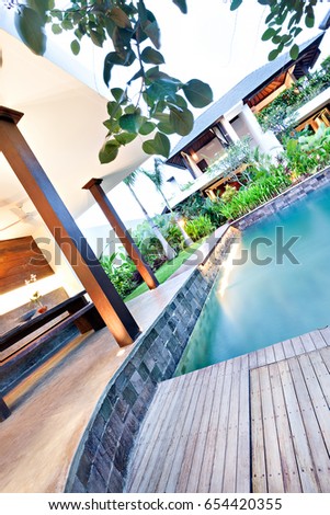 Modern swimming pool and wooden sidewalk around in a luxury house with pillars in front of the garden and the house, looking under the branches and leaves