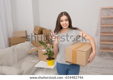 Pretty young woman standing with cardboard box in her hand showing ok with her fingers. Female in the living room ready to unpack boxes after relocating.