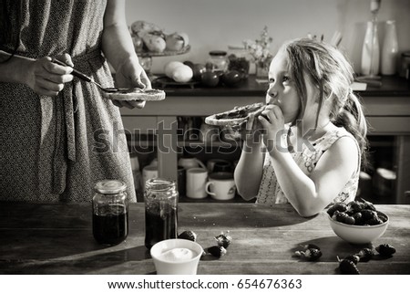 A mom preparing a slice of strawberry jam for her little girl in the kitchen. There are strawberries and sugar on the table. Black and white picture