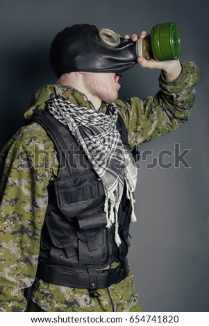 A soldier in camouflage and bulletproof vests takes off a gas mask. A man is gasping for a gas attack. Studio photo on a gray background.