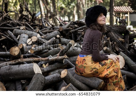 Attractive middle aged woman with black fur Russian winter hat, long sleeve brown shirt and orange pants sitting on firewood in the park. Mature lady model outdoors. Fall fashion sales concept