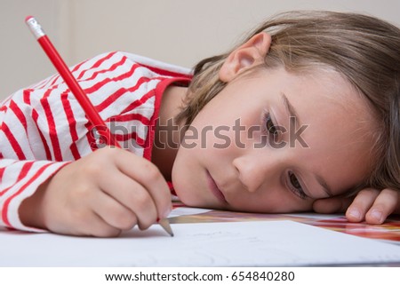 A portrait of a tired little girl writing with a red pencil on a white paper sheet