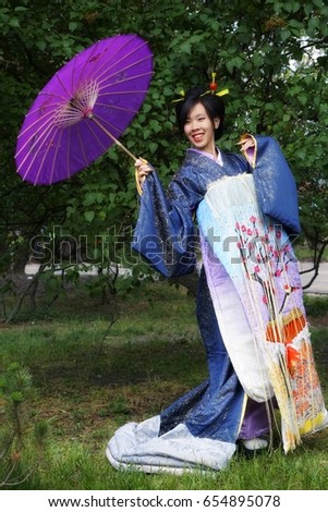 Smiling happy japanese woman of appearance with black hair in national Japanese costume with violet umbrella. Hair is assembled in hairstyle with deco sticks. Long blue dress with japanese design Royalty-Free Stock Photo #654895078