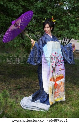 Smiling lucky japanese girl of appearance with black hair in national Japanese costume with violet umbrella. Hair is assembled in headdress with deco sticks. Long blue dress with japanese  tracery Royalty-Free Stock Photo #654895081