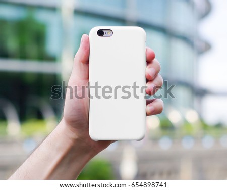 Smart phone on a blurry city background. in a white plastic case back view. Smart phone in man's hand. Template of smart phone case. Mock up of phone case Royalty-Free Stock Photo #654898741