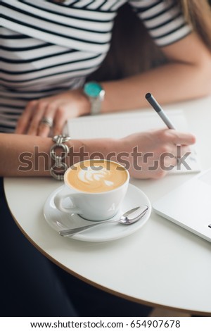 Close-up picture of hands making notes on paper, cup of cappucciono and laptop standing on a table in a cafe.