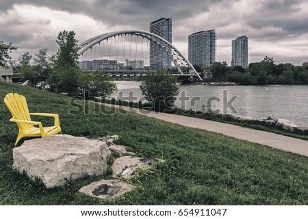 Yellow wooden chair by the water on a dark overcast day with arch bridge and buildings in the background.