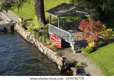 Luxury lake waterfront rocky beach property with covered wood deck patio sitting area next to water in landscaped backyard garden beside stack of plastic kayak recreation boats in Washington dusk sun