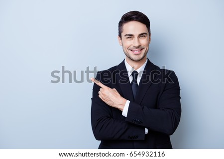 Close up portrait of young successful brunete  stock-market broker guy on the pure light blue background, he is smiling, wearing suit with tie and is pointing on a copyspace with his finger