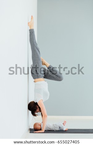 Vertical photo of sporty mother on hands upside down while her little son lying near her on mat in home. Gorgeous mom with her cute baby having fun together. Fitness, maternity, healthy life concept.