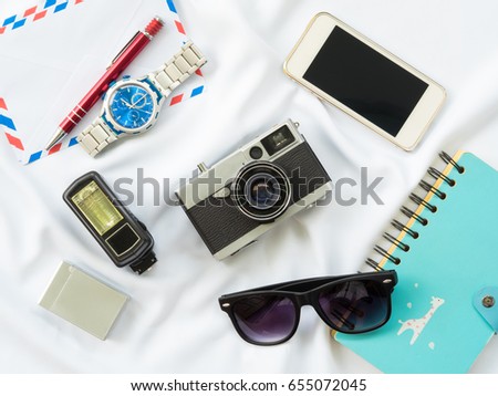 Flat Lay photo with prop are Air letter, watch, pen, Flash, Camera, Glasses, Charger, Blue Notebook and smartphone on white fabric background.