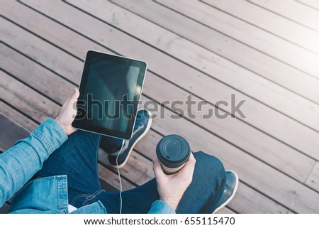 View from above. Close-up of tablet computer with black blank screen and cup of coffee in hands of hipster man sitting on bench outside.Mock up.Space for logo, text, image. Guy chatting, blogging.