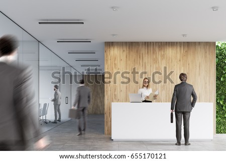 Rear view of businessmen near a white reception desk with two laptops standing on it in front of a wooden office wall. There are glass wall offices to the left. Royalty-Free Stock Photo #655170121