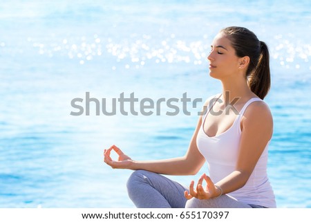 Close up portrait of young woman meditating in yoga position outdoors. Girl in casual wear sitting against blue sea background with sun reflecting on water surface.