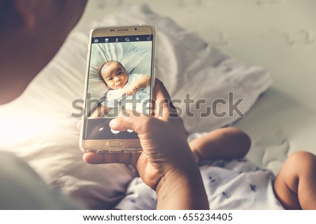 Above high angle shot of happy mom taking photo her Asian baby boy in bed with a smartphone at home. It is a memorable authentic feeling for every parent when their child is growing up. Top down view