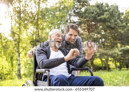 Hipster son and father in wheelchair at park taking selfie.