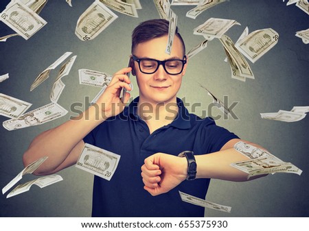 Busy businessman looking at wrist watch, talking on mobile phone under cash rain. Time is money concept  